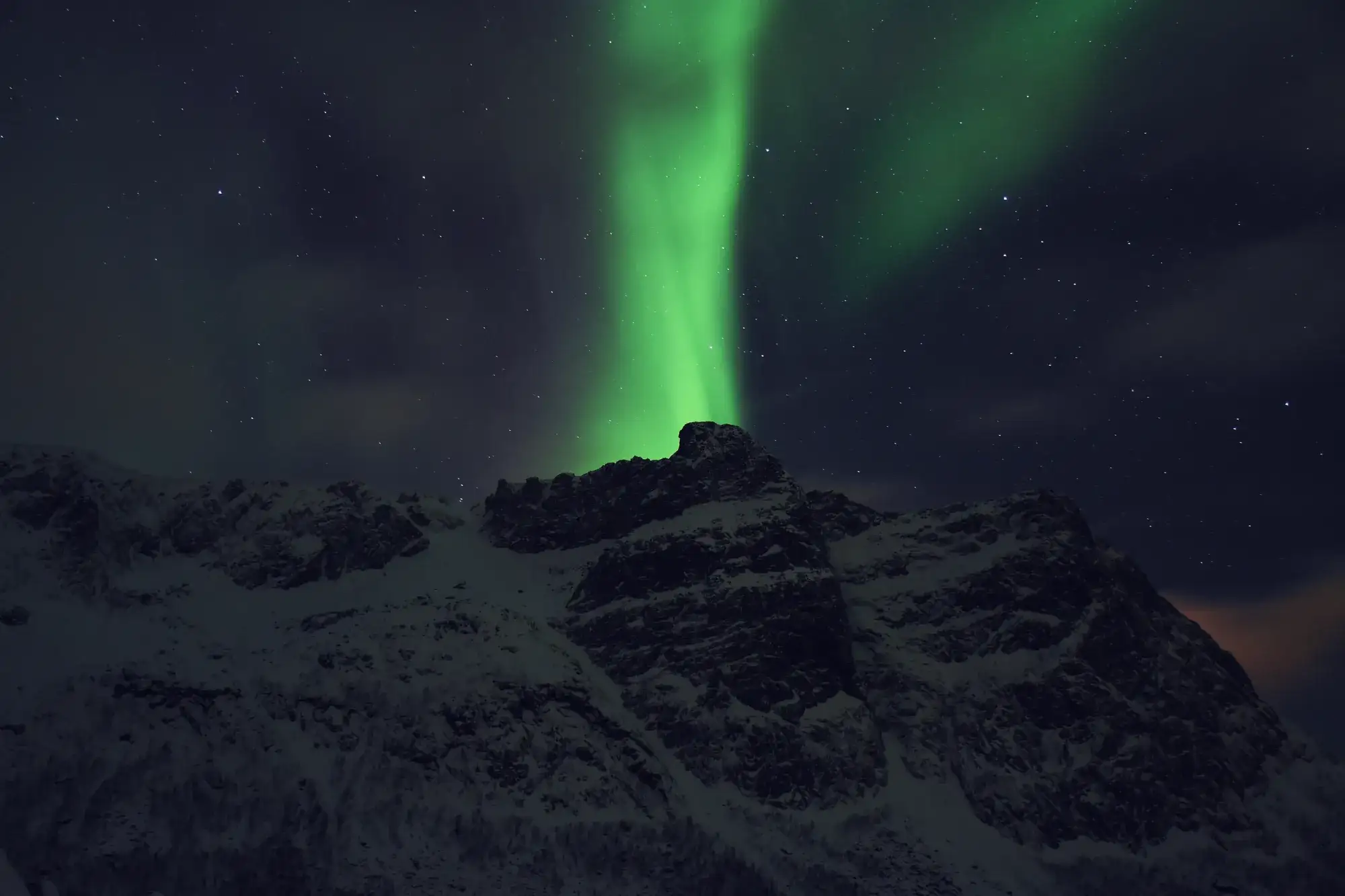 A green aurora pillar rises above snow-covered mountains near Tromsø, Norway.